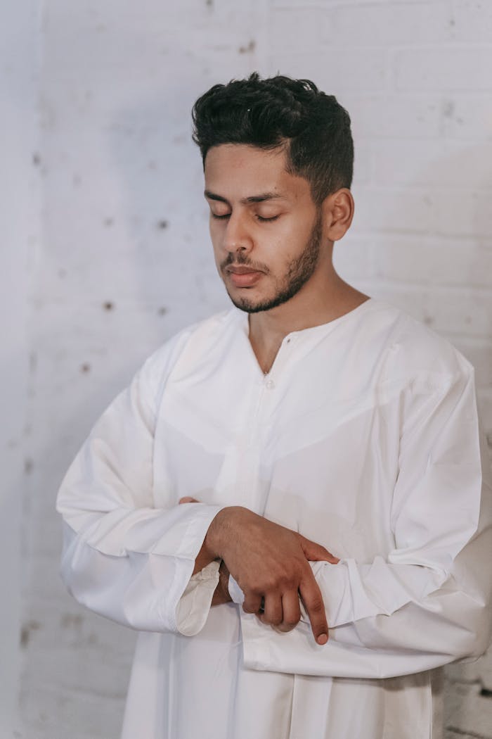 A man in a white thobe stands in prayer position indoors, embodying Islamic faith.