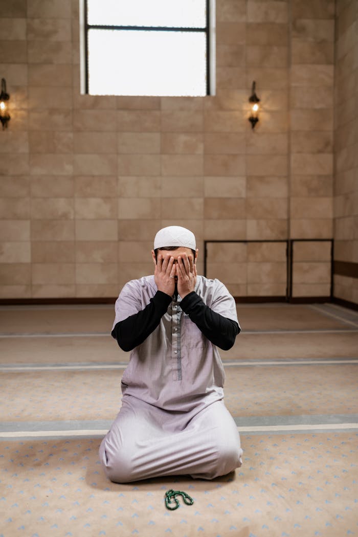 A man kneeling and praying inside a mosque, hands covering face, reflecting deep faith.